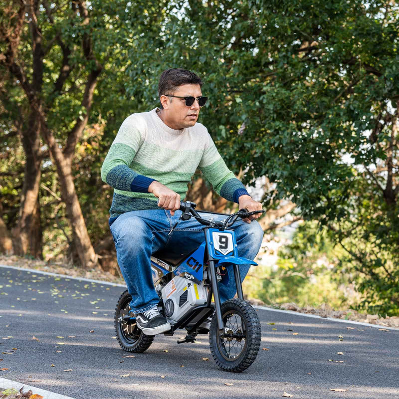 A rider on a blue KerryYoo electric dirt bike for kids on a tree-lined trail